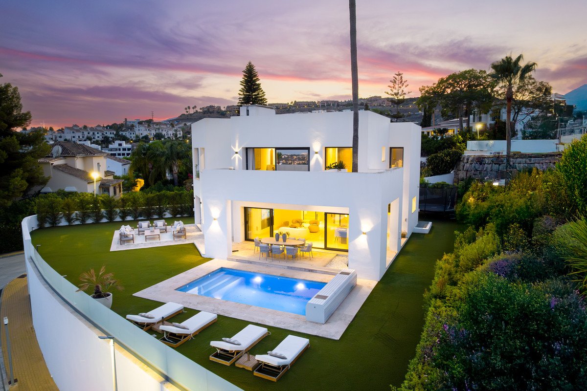 Modern white villa with a swimming pool and lounge chairs, overlooking a town and hills at sunset.