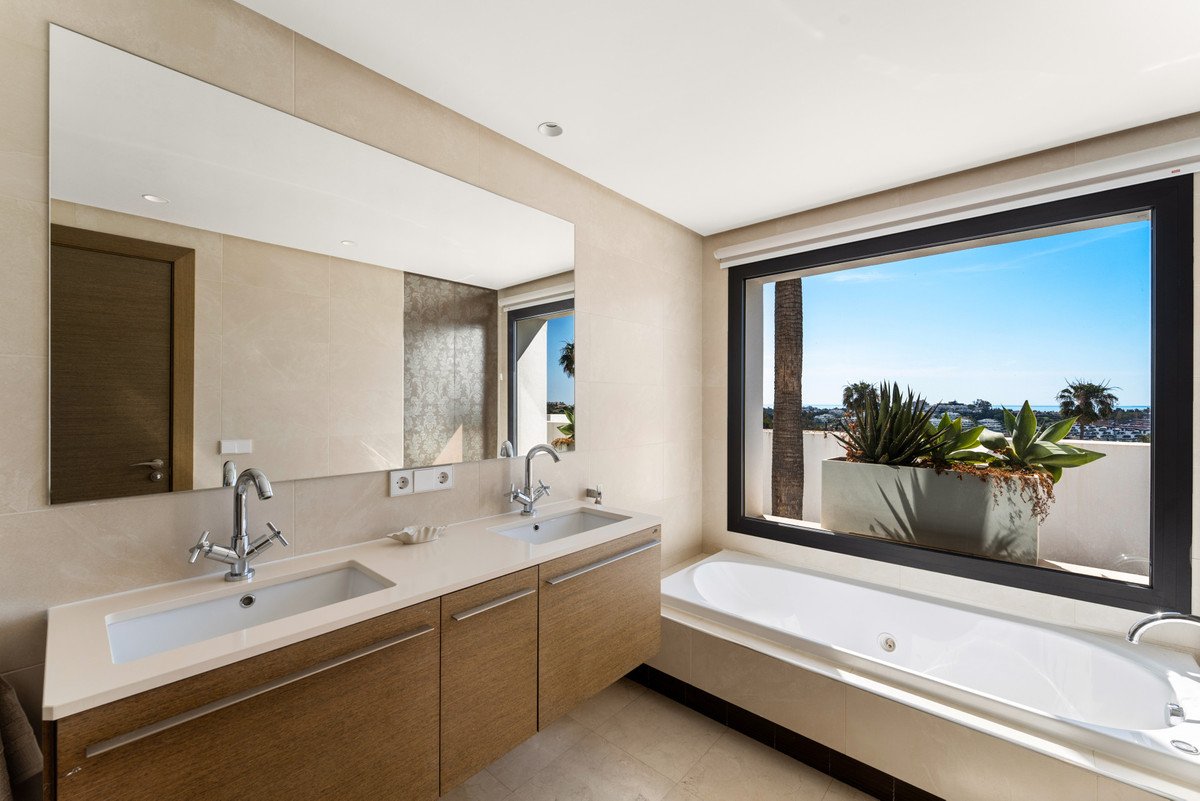 Modern bathroom with dual sinks, a bathtub, and a large window offering an ocean view and lush greenery outside.