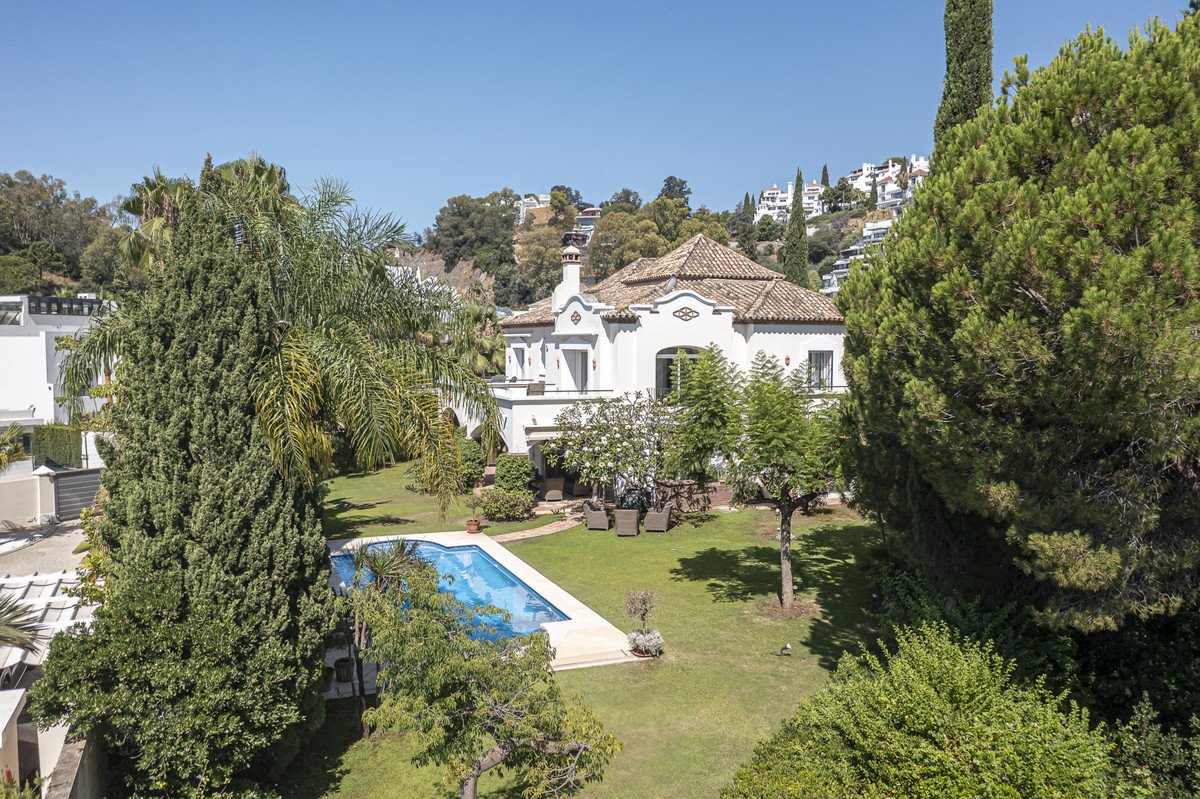 A white villa with a swimming pool and garden, set against a backdrop of trees and hills.