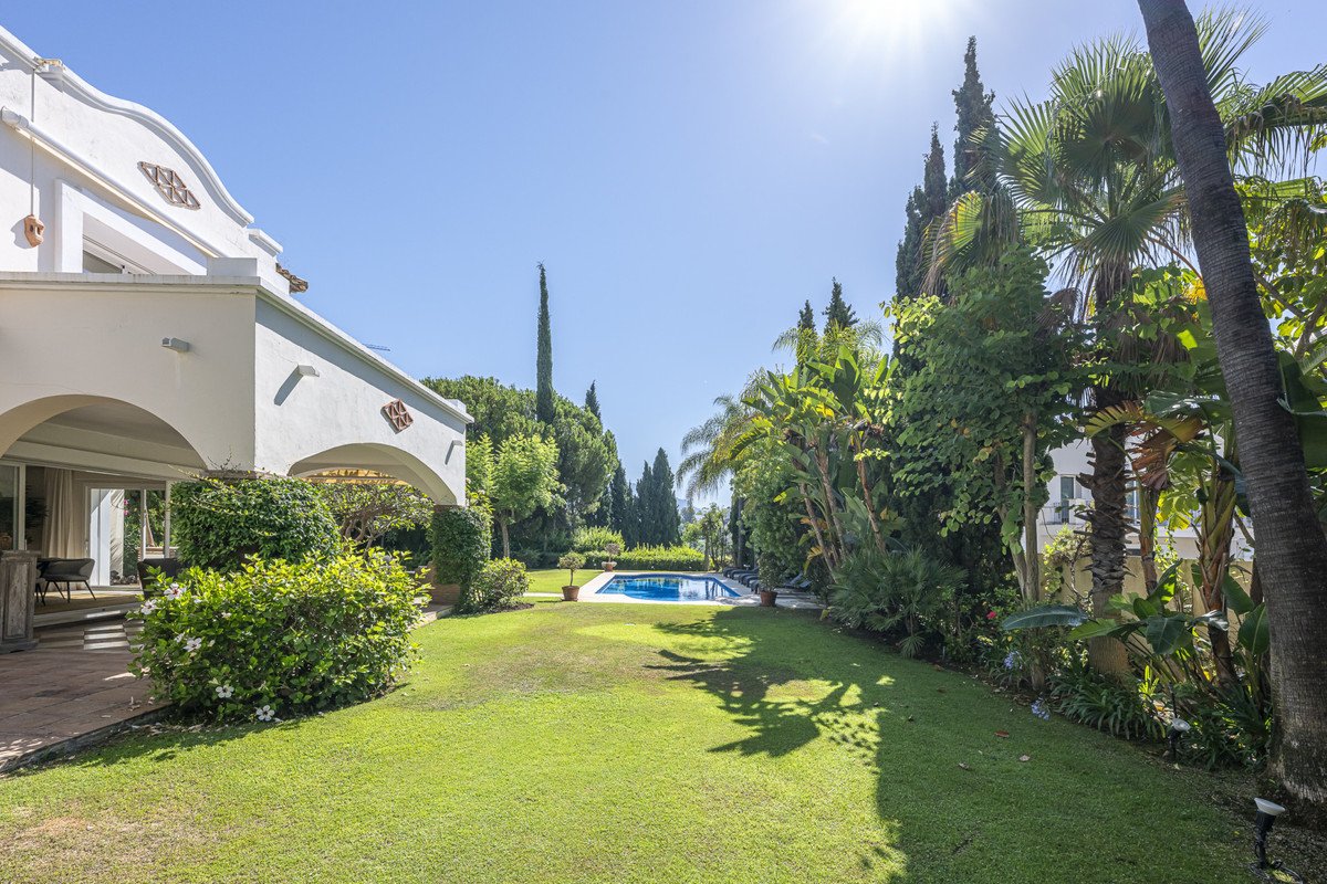 A white villa with arches opens onto a lush green lawn and a bright blue swimming pool surrounded by mature trees.