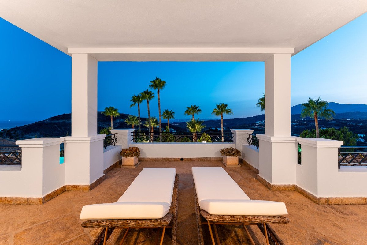 Spacious terrace with loungers, offering panoramic views of the sea, hills, and palm trees at dusk.