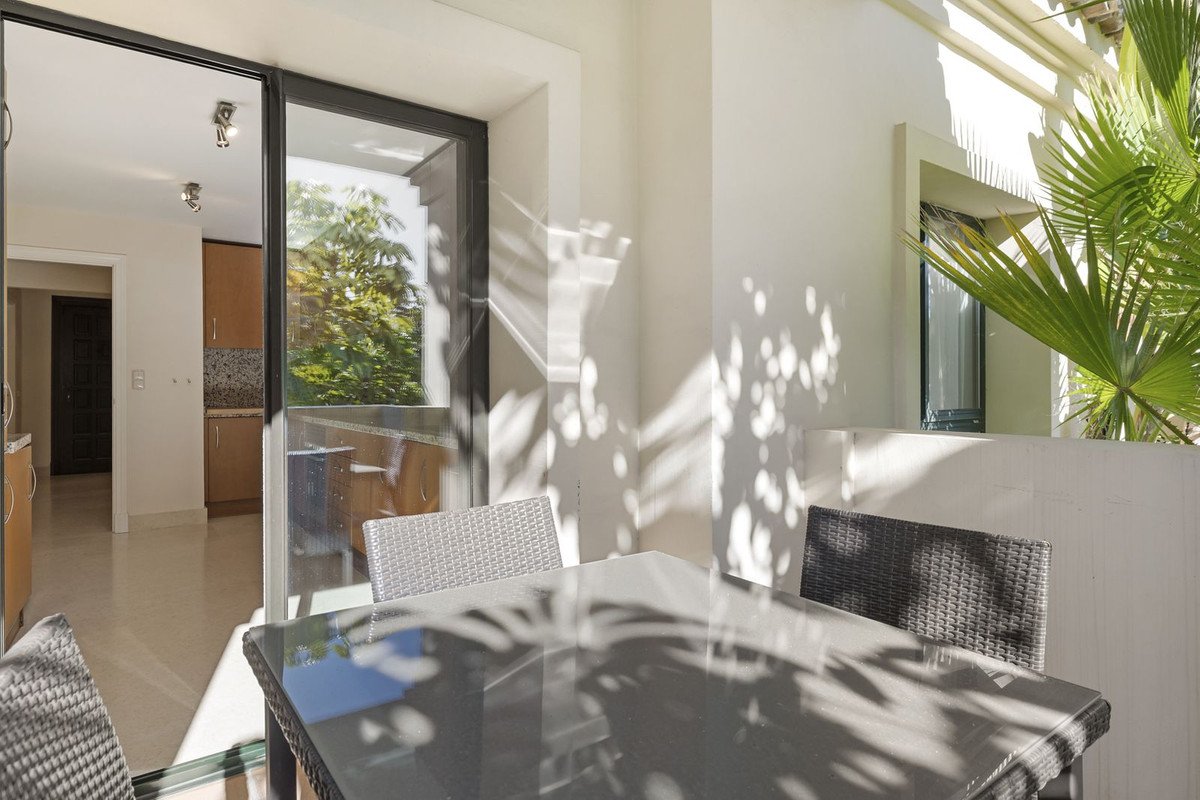 Terrace dining area with chairs and table, visible kitchen through sliding glass doors, lush green trees outside.