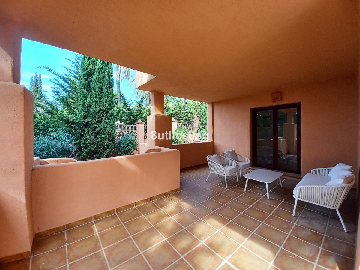 Covered terrace with garden view and outdoor seating area, featuring white wicker furniture and a glass door leading inside.
