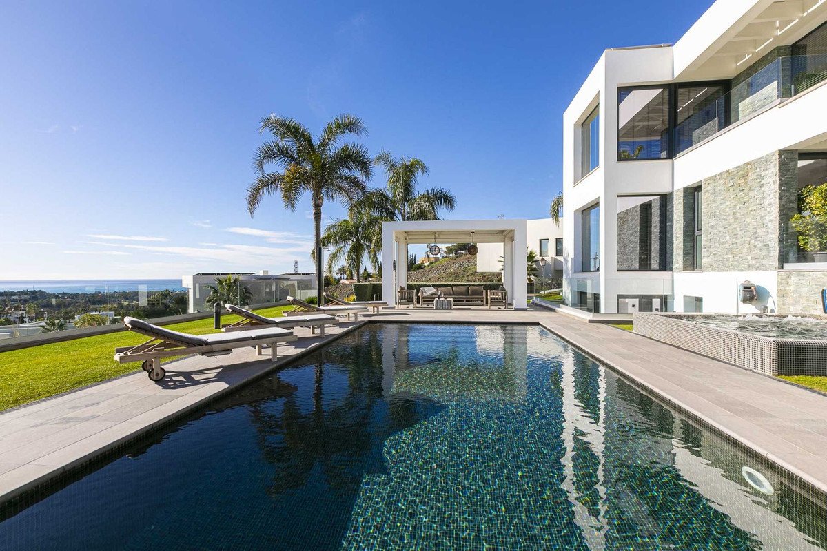 A modern villa with an infinity pool, lounge chairs, and a covered outdoor seating area, overlooking the sea.