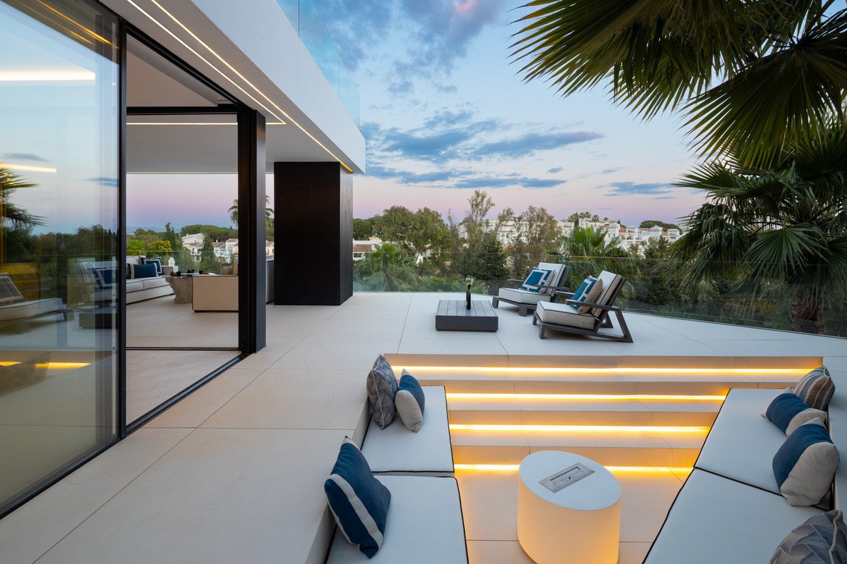 Modern terrace with built-in seating, lounge chairs, and a coffee table overlooking a lush green landscape at dusk.