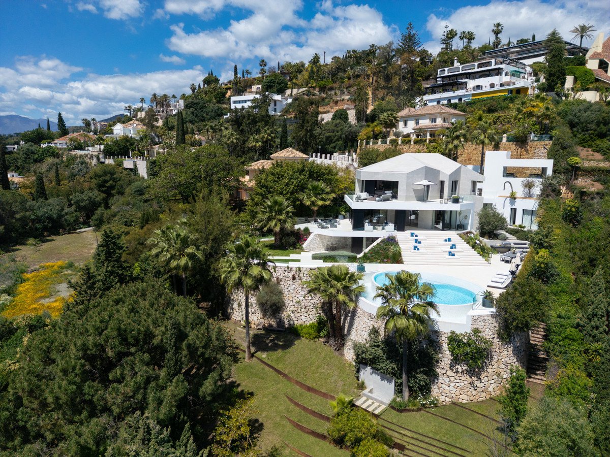 Modern white villa with swimming pool and lounge area on a sunny day with lush green surroundings and a distant sea view.