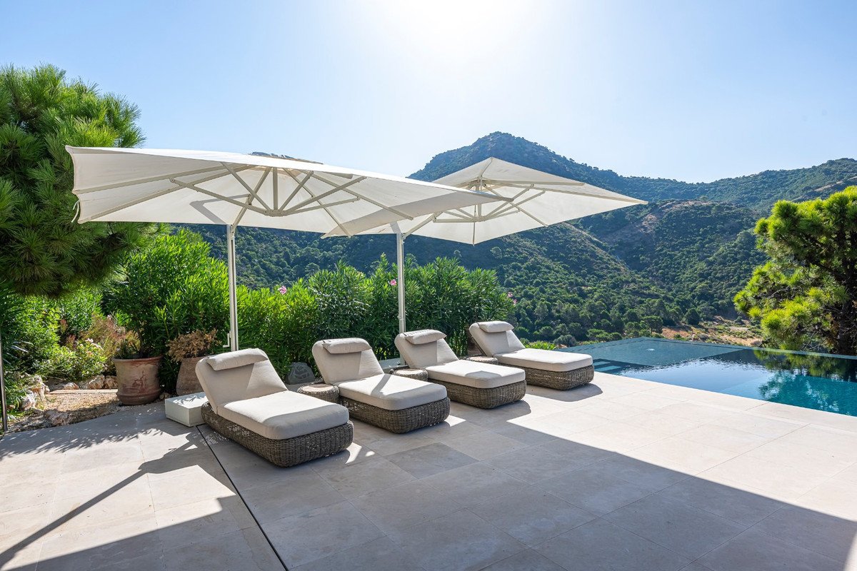 Four lounge chairs with umbrellas beside an infinity pool overlooking a lush mountain landscape on a sunny day.