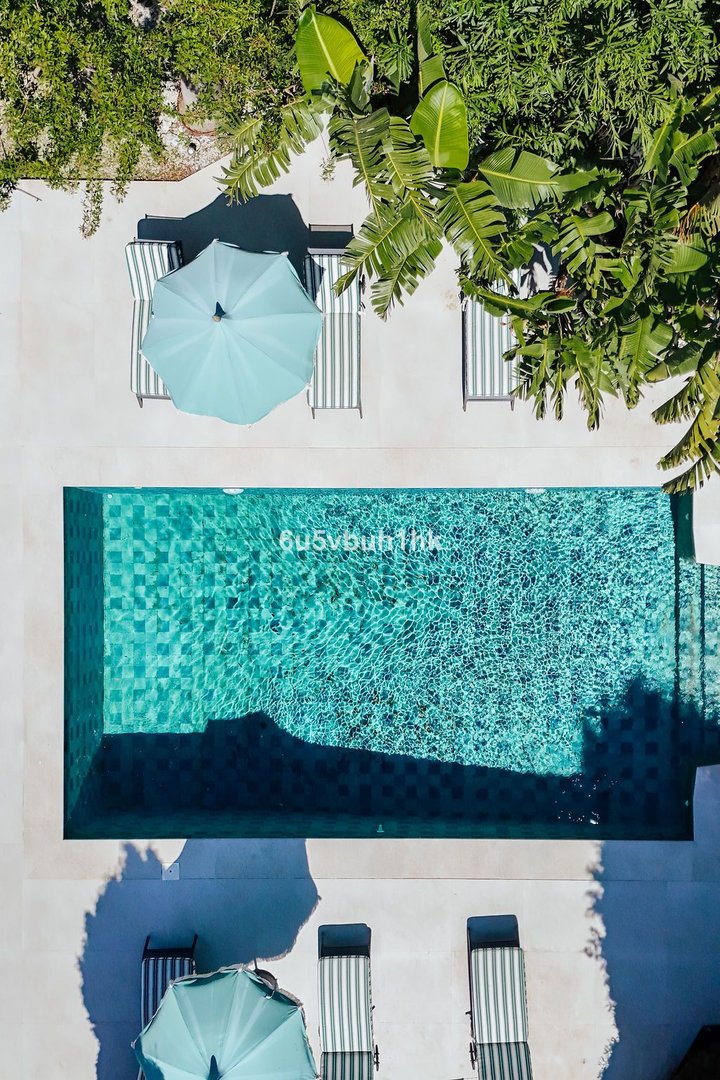 Overhead view of a turquoise swimming pool with lounge chairs and umbrellas, surrounded by lush greenery and shadows.