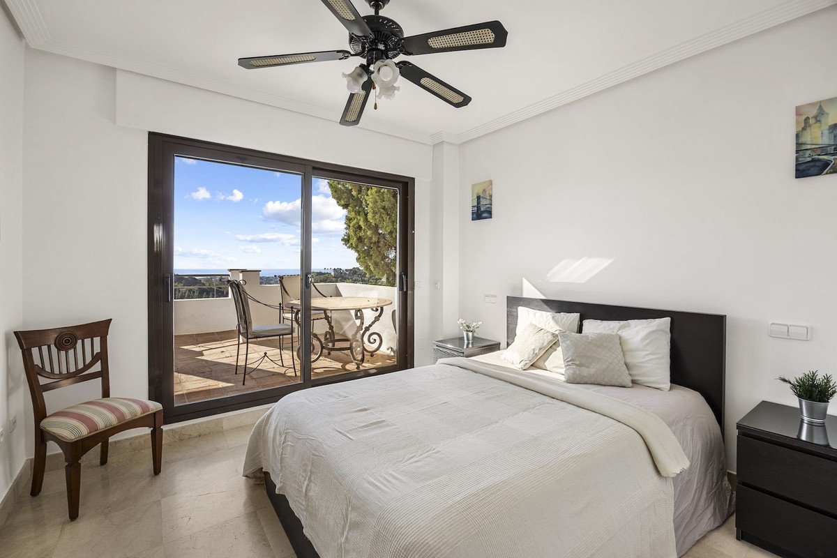 Bedroom with a large bed, a ceiling fan, and a sliding glass door leading to a balcony with a sea view and a table and chairs.