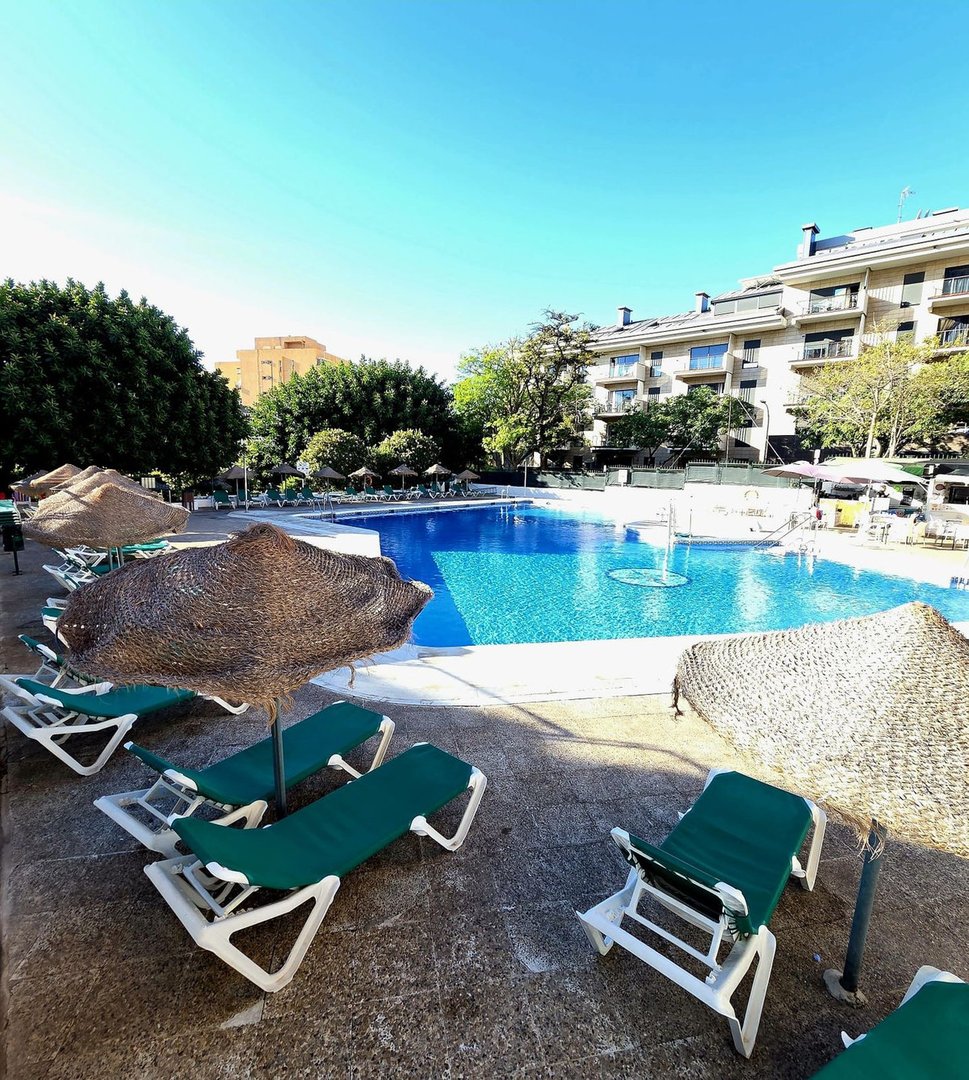 Green loungers and thatched umbrellas surround a large blue swimming pool with a building in the background under a clear blue sky.