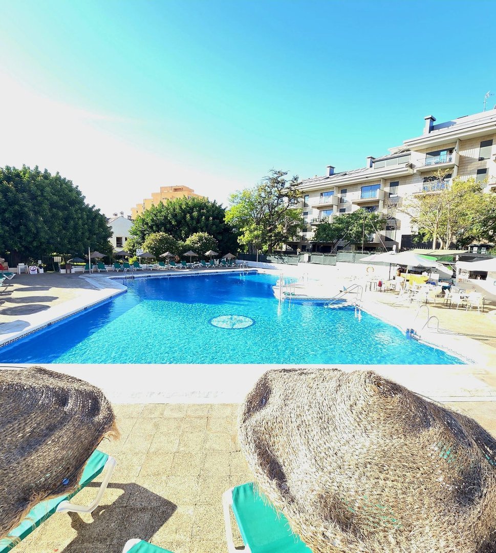 Large sparkling blue swimming pool with lounge chairs and thatched umbrellas, surrounded by trees and buildings.