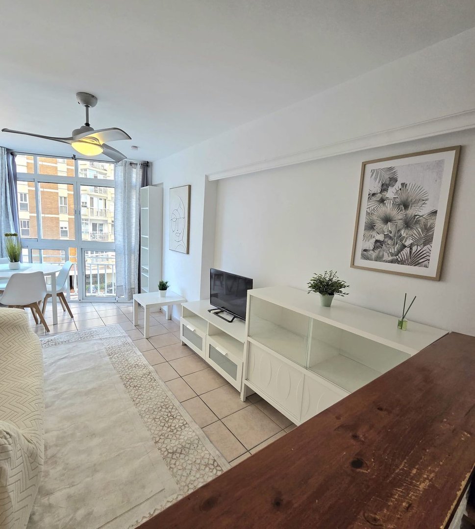 Modern living room with city view, featuring a TV, white furniture, a dining area, and large windows with curtains.