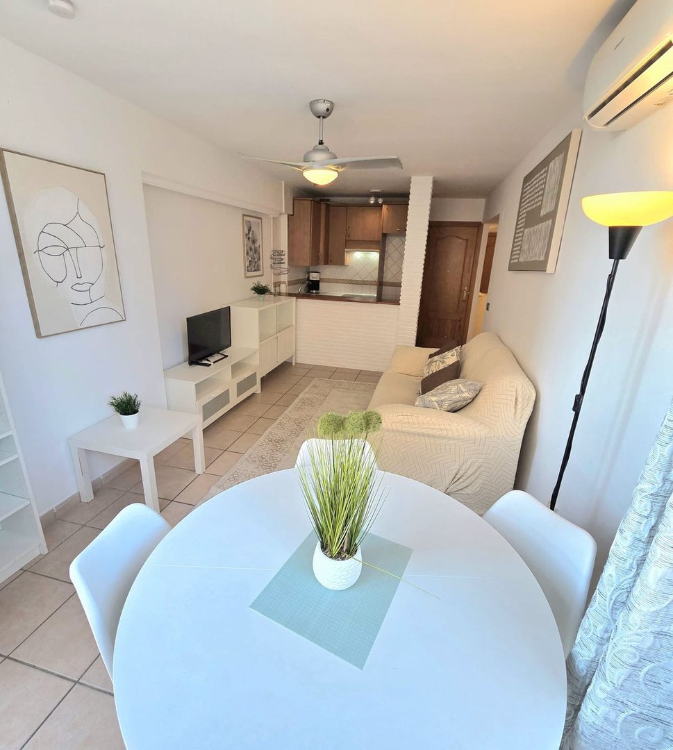 A bright living area with a sofa, dining table, and kitchenette, featuring modern decor and a ceiling fan.