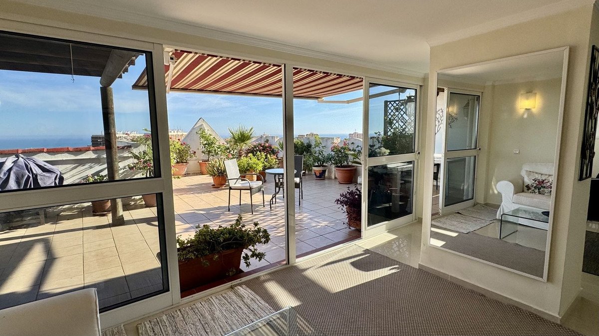 A bright living room opens onto a spacious terrace with potted plants, seating, and a distant sea view under a striped awning.