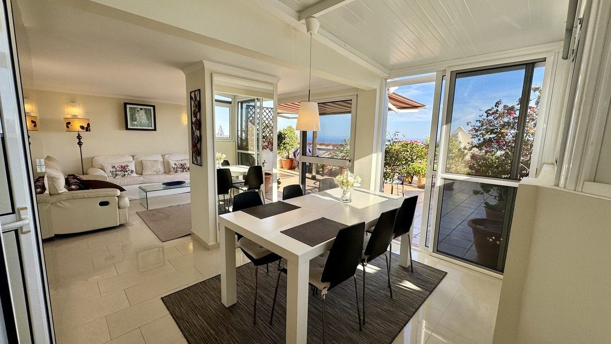 A bright dining area with a white table and chairs, leading to a sunroom with large windows overlooking the sea and greenery.