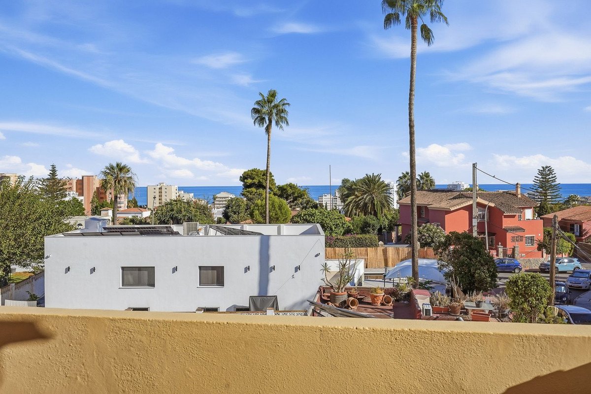 View from a rooftop balcony with palm trees, residential buildings, and the blue sea in the distance under a clear sky.
