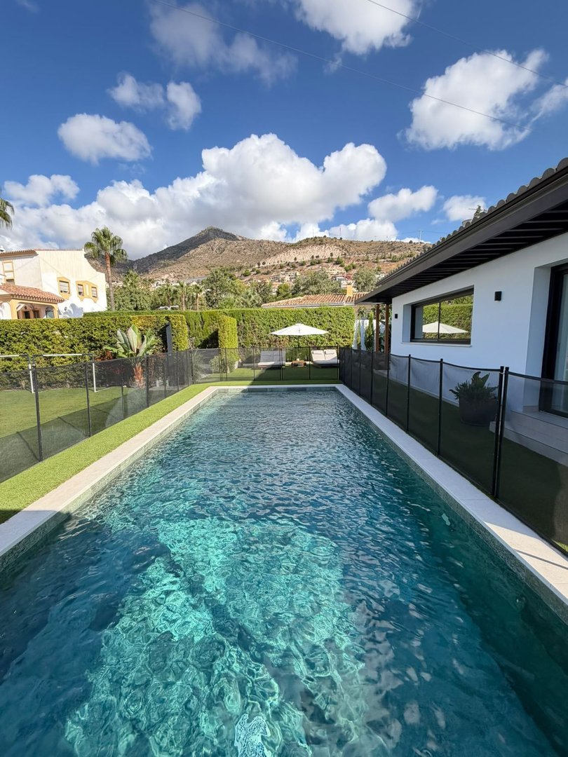 A long swimming pool with clear blue water, surrounded by a green lawn and a fence, with houses and mountains in the background.