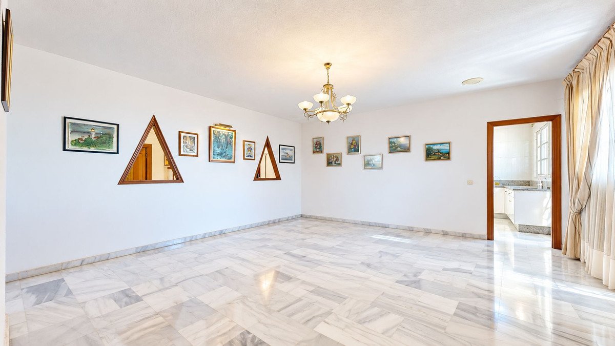 A spacious living room with white walls, marble floors, a chandelier, and several framed artworks on the walls.