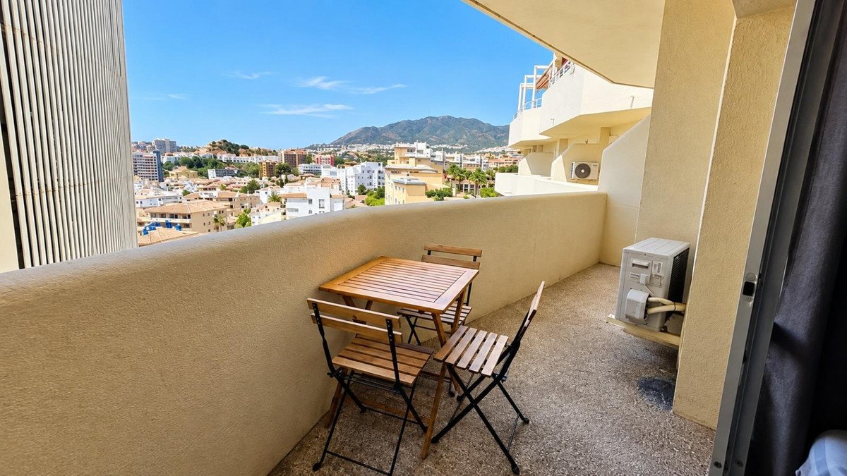 Balcony with a small wooden table and chairs, overlooking a town and mountains under a clear blue sky.