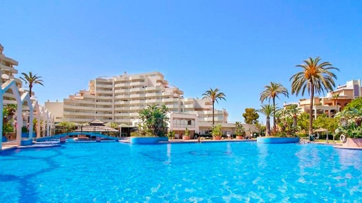 A large, bright blue swimming pool with palm trees and apartment buildings in the background under a clear blue sky.