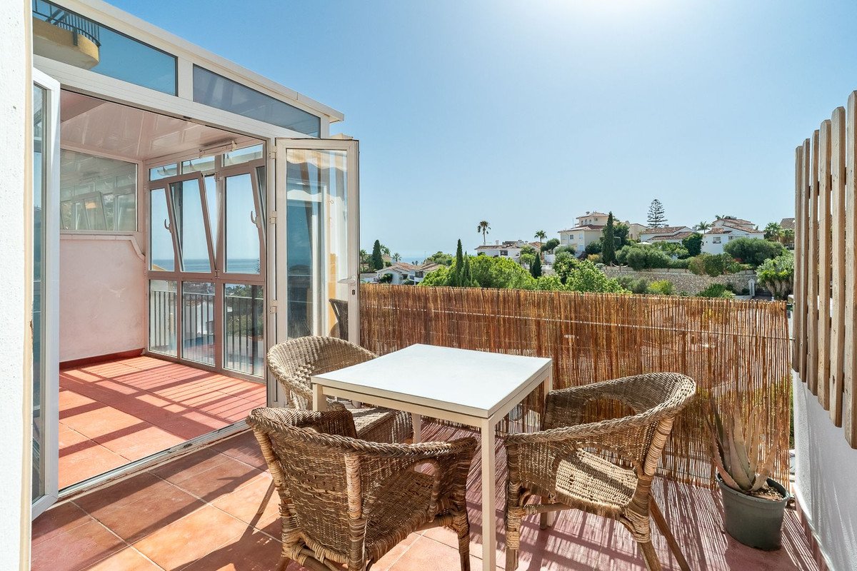 A sunlit terrace with wicker chairs and a table overlooking a sea view and distant hills with houses.