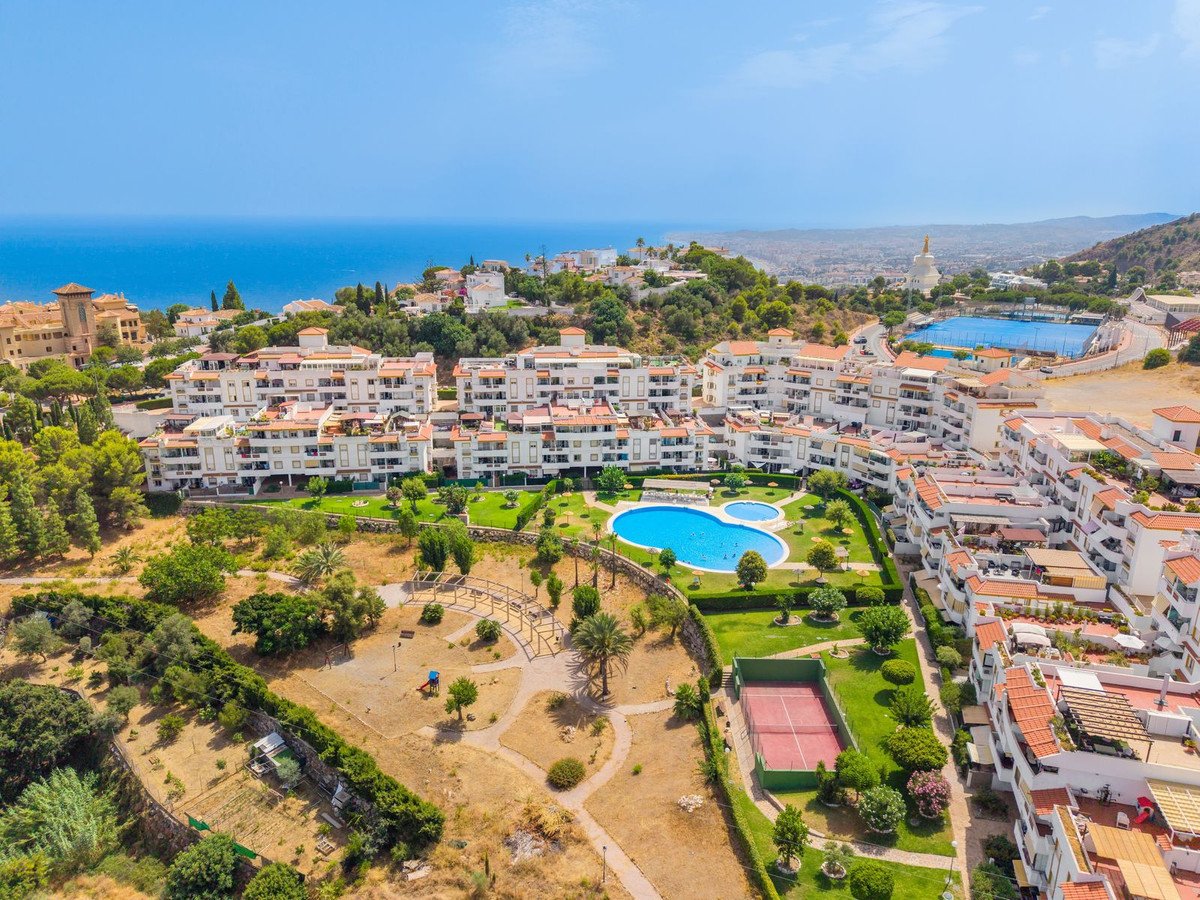 Aerial view of a Mediterranean apartment complex with a large swimming pool, tennis court, and distant sea views.