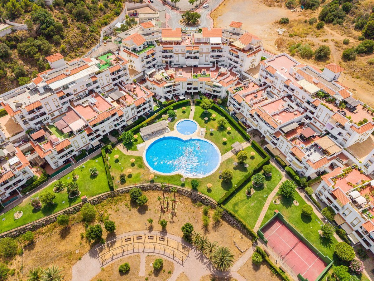 Aerial view of resort apartments with a large swimming pool, manicured gardens, and a tennis court on a sunny day.