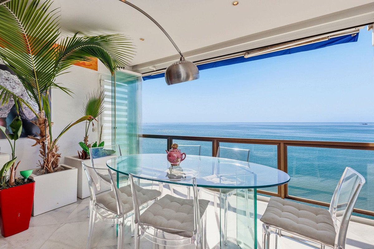 Glass dining table and chairs on a balcony with a stunning sea view and lush greenery.