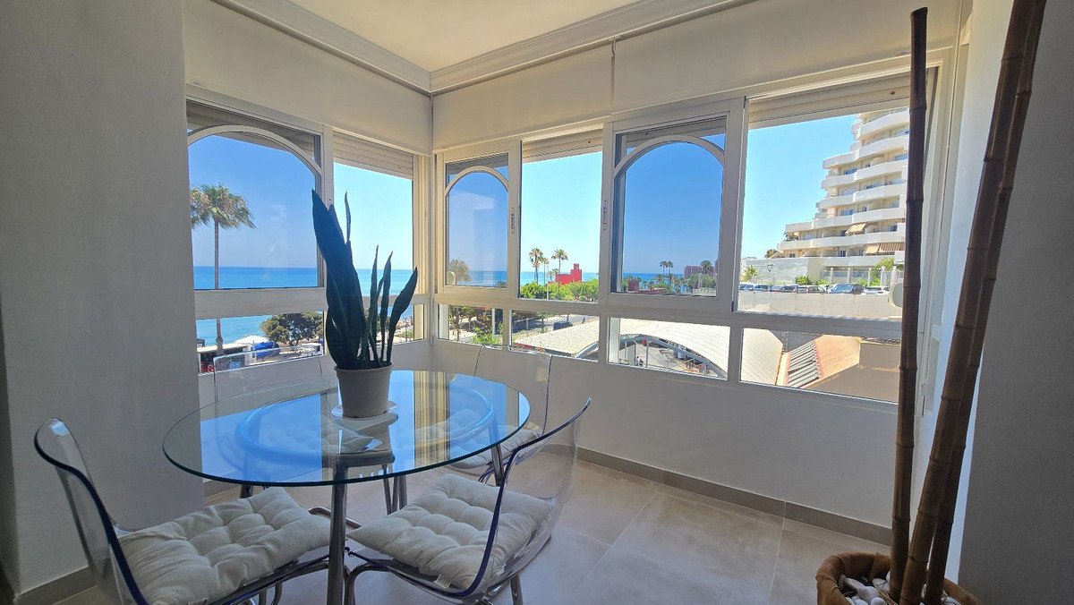 Dining area with a glass table and chairs, featuring a large window with a view of the sea and palm trees.