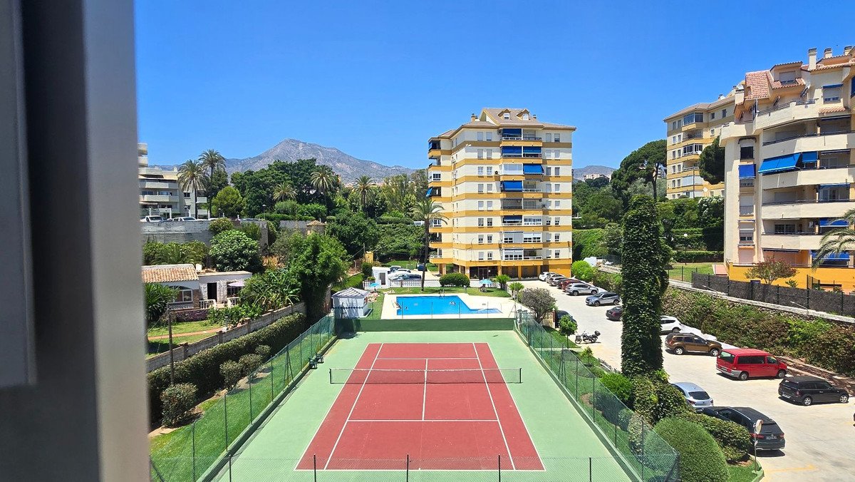 A view overlooking a red tennis court and blue swimming pool with apartment buildings and mountains in the background.