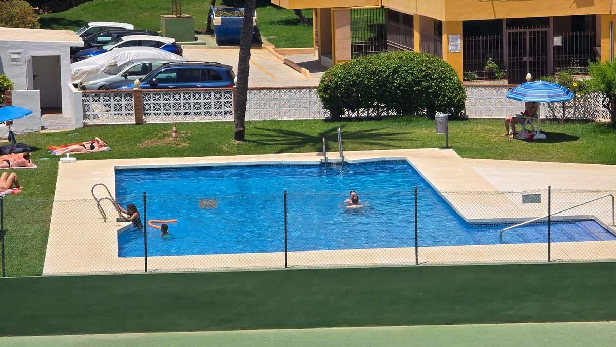 A vibrant blue swimming pool with people relaxing and sunbathing on the grassy lawn under a clear sky.