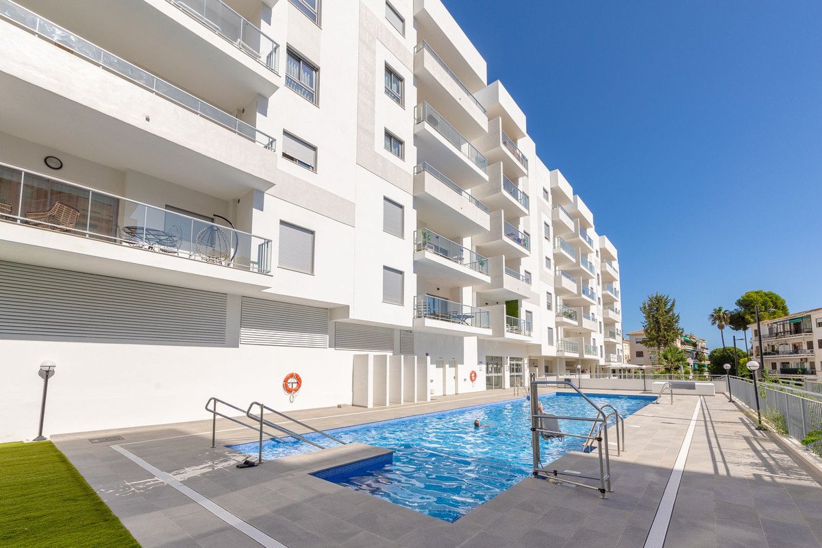 Exterior of a modern white apartment building with balconies and a bright blue swimming pool on a sunny day.