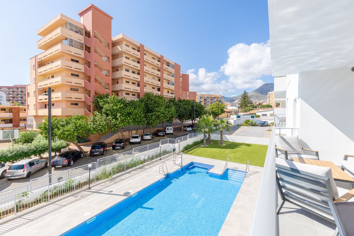 A balcony overlooking a swimming pool, green lawn, and apartment buildings with mountains in the background under a blue sky.