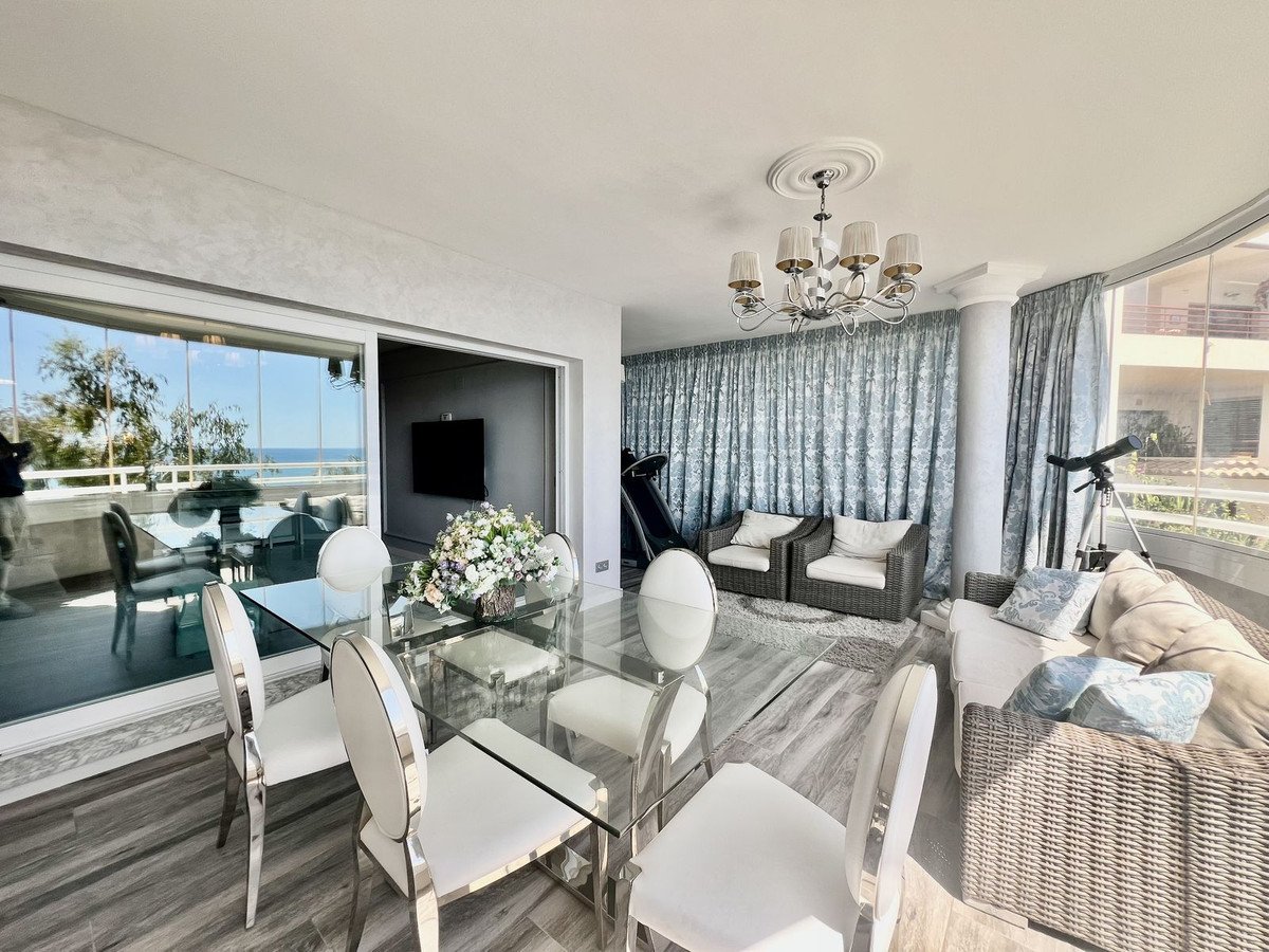 Spacious living and dining area with a glass table, elegant chairs, and a view of the sea through large windows.