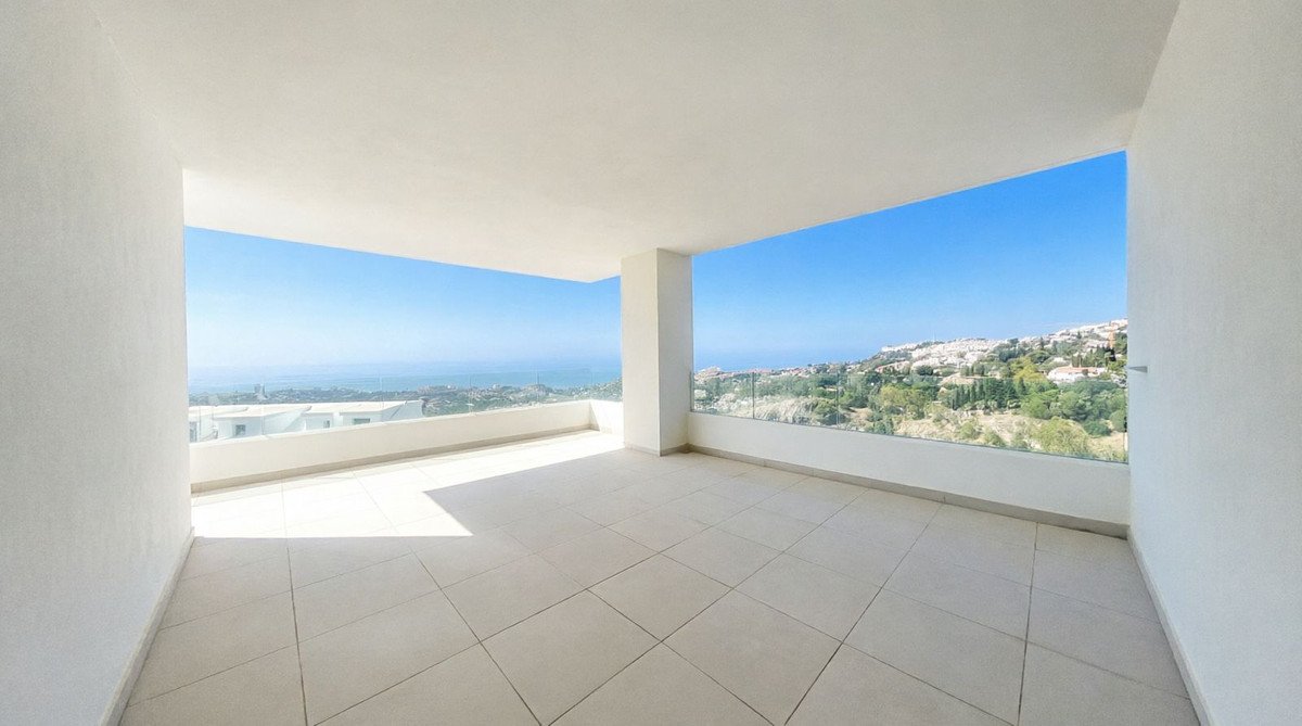 Spacious balcony with panoramic views of the sea, hillside town, and clear blue sky. Modern glass railings and tiled floor.