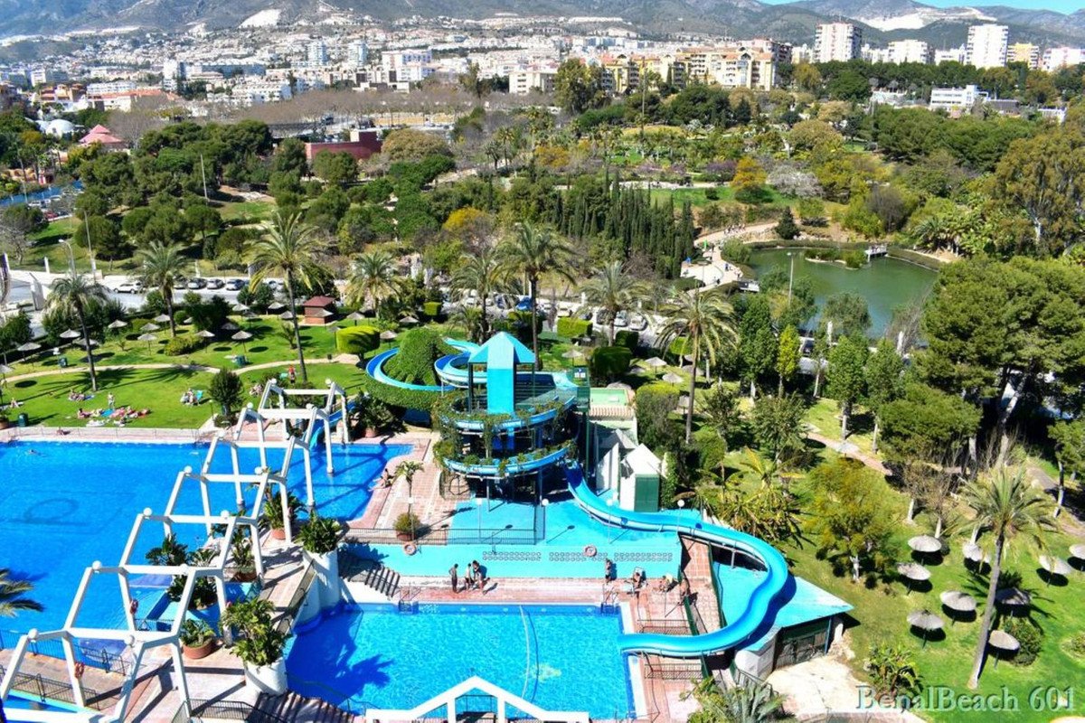 Aerial view of a water park with blue pools, slides, tropical gardens, and city in the background.