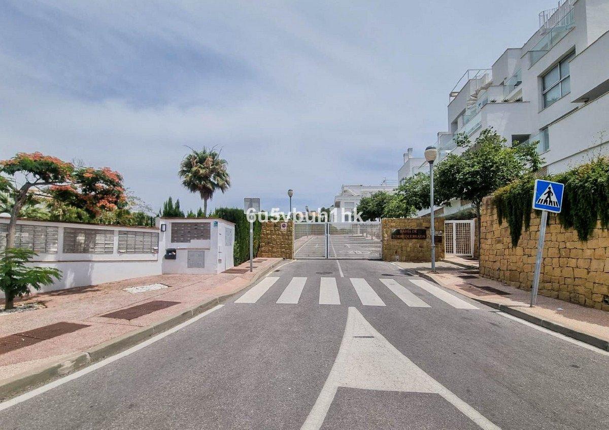 Modern apartment complex entrance with gated access, palm trees, and pedestrian crossing under clear sky.