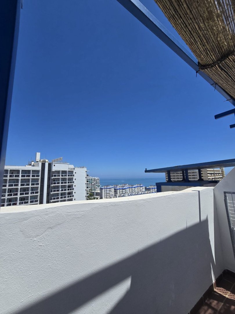 Balcony view overlooking buildings and the sea under a clear blue sky with a thatched roof overhead.