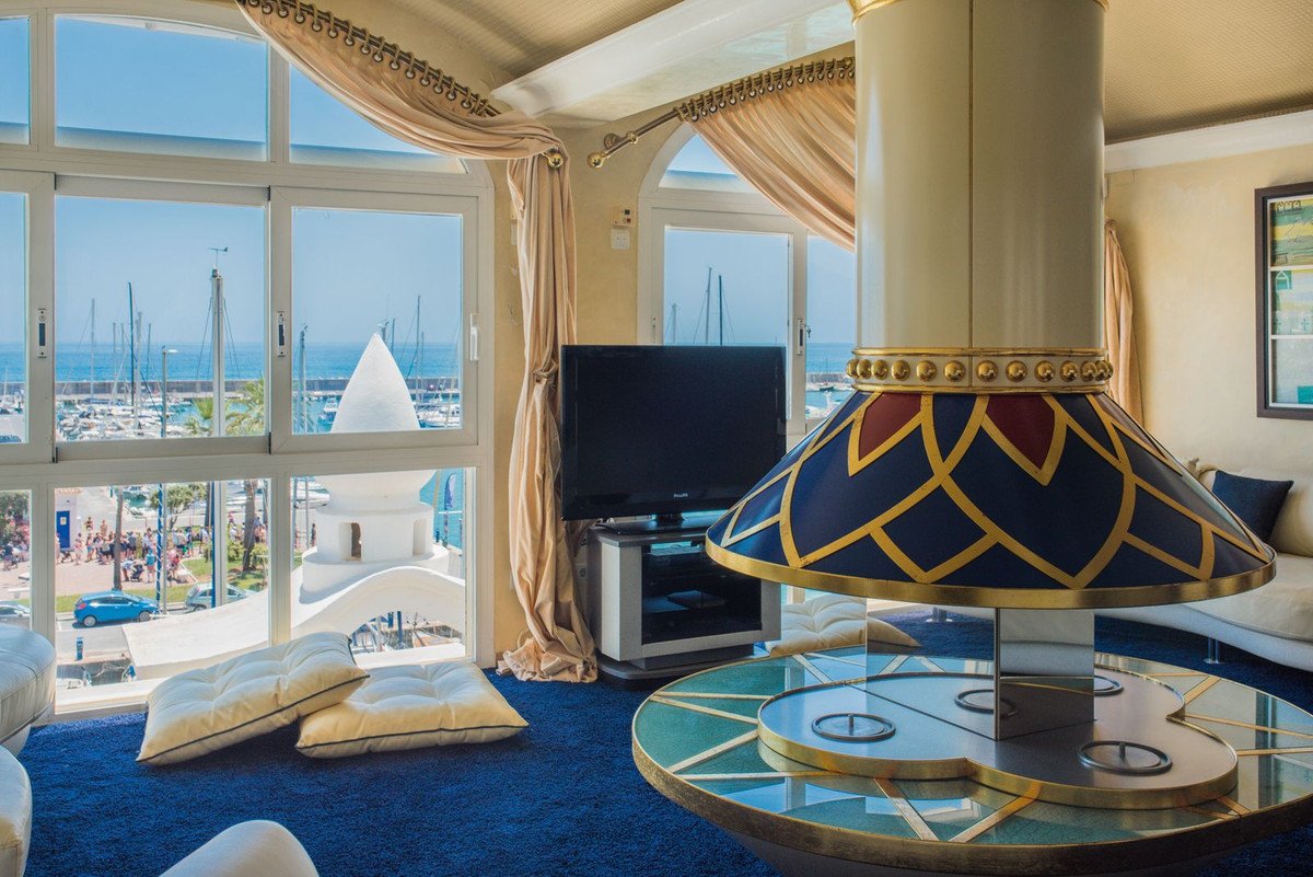 Living room with large windows overlooking a marina and the sea, featuring a decorative fireplace and blue carpet.