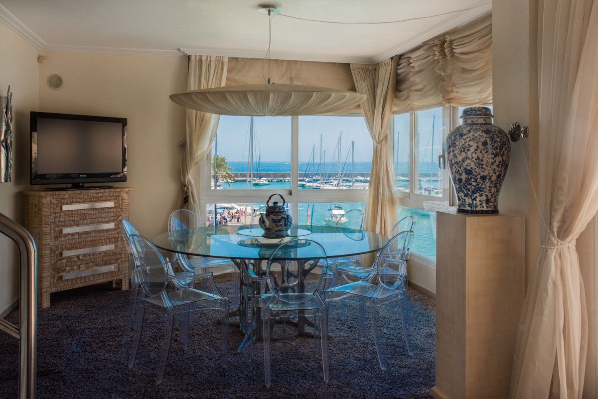 Dining area with glass table and chairs overlooking a marina filled with yachts and the blue sea.
