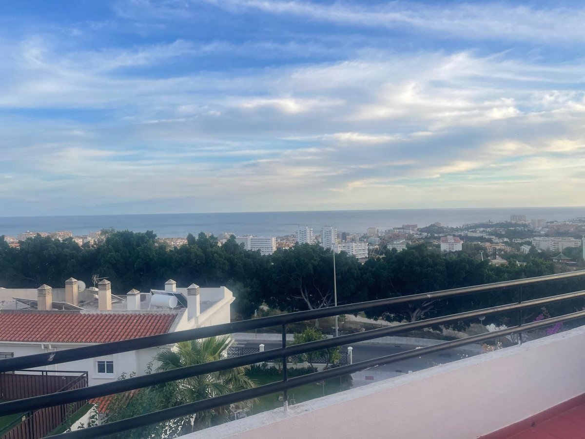 A scenic view from a balcony overlooking the sea and a coastal town, with trees in the foreground.