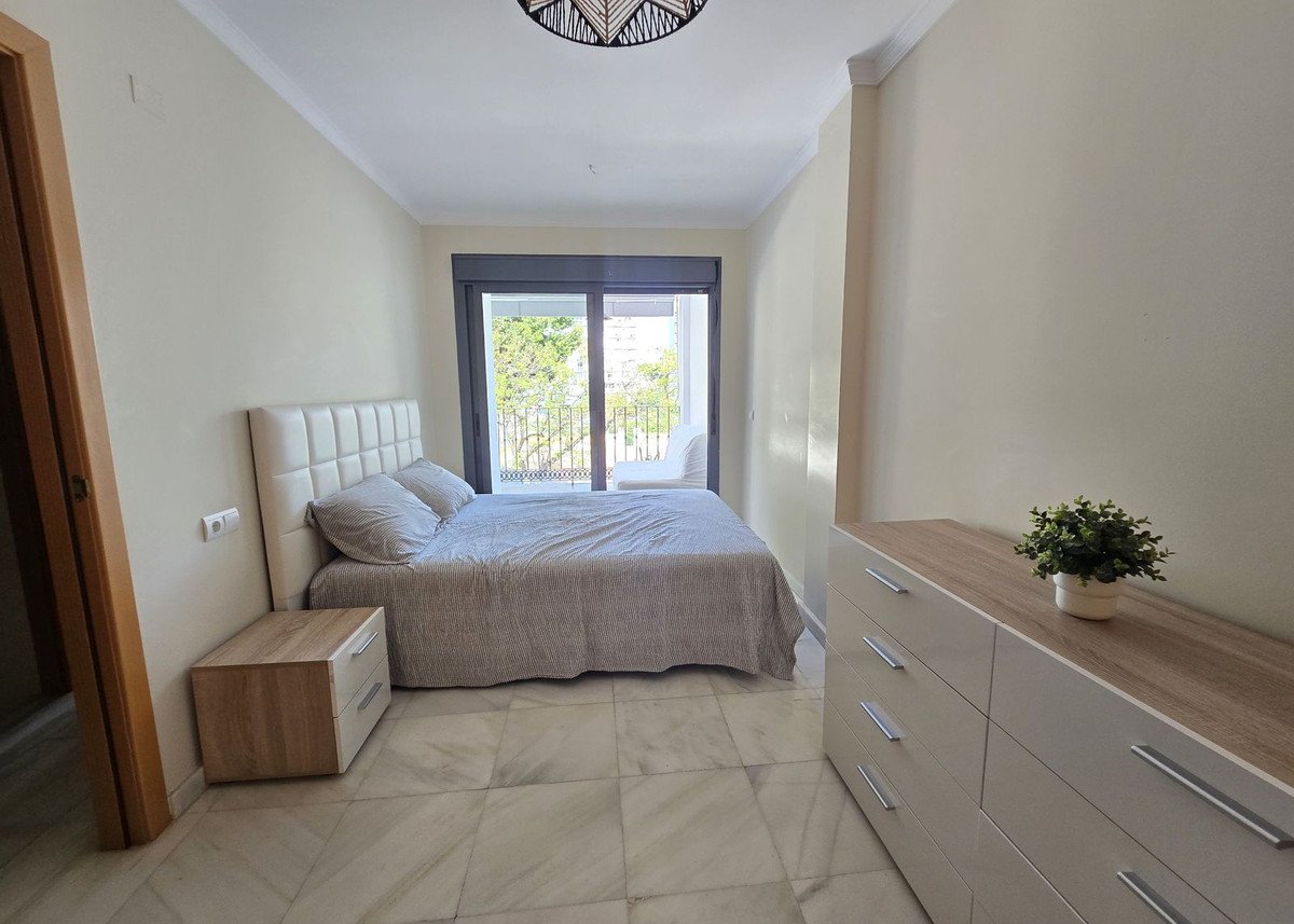 A bedroom with a queen-size bed, nightstand, dresser, and balcony access with a view of trees and buildings.