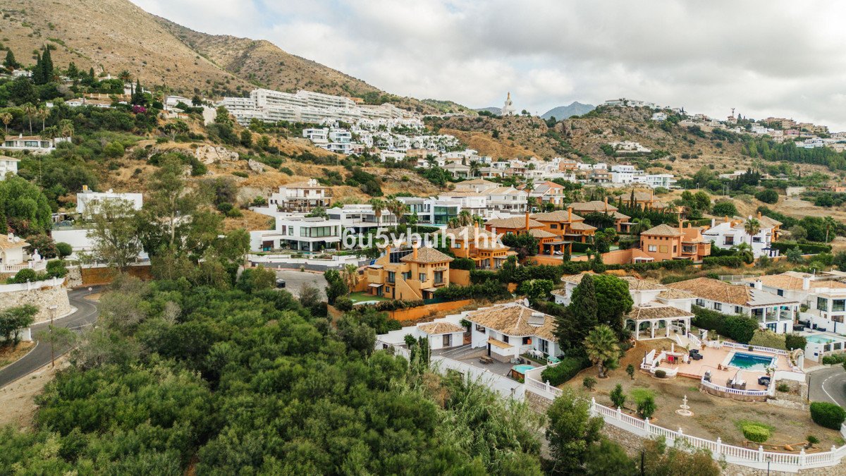 Panoramic view of hillside villas with a swimming pool and lush greenery under a cloudy sky.