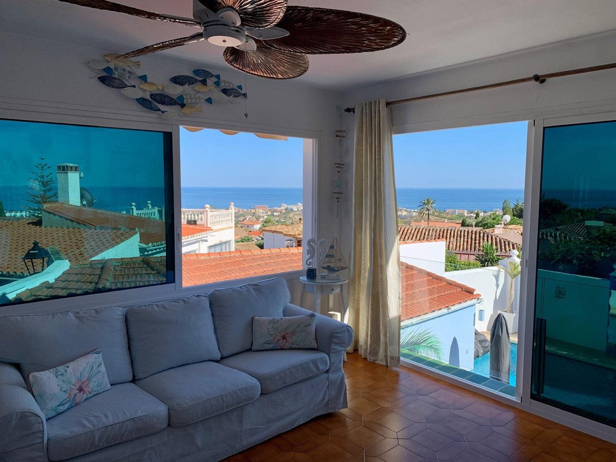 Living room with a sofa and large windows offering a panoramic view of the sea, rooftops, and a swimming pool.