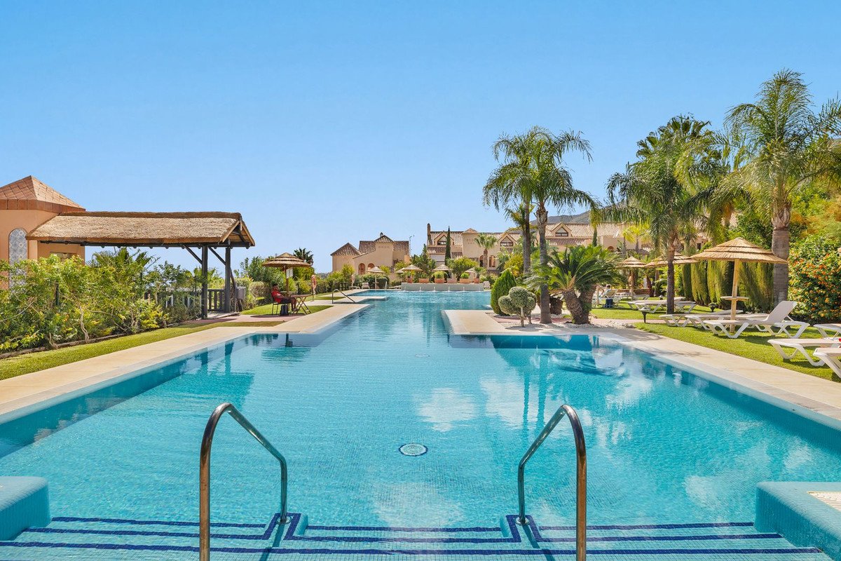 A long, clear blue swimming pool with palm trees and resort buildings in the background under a clear sky.