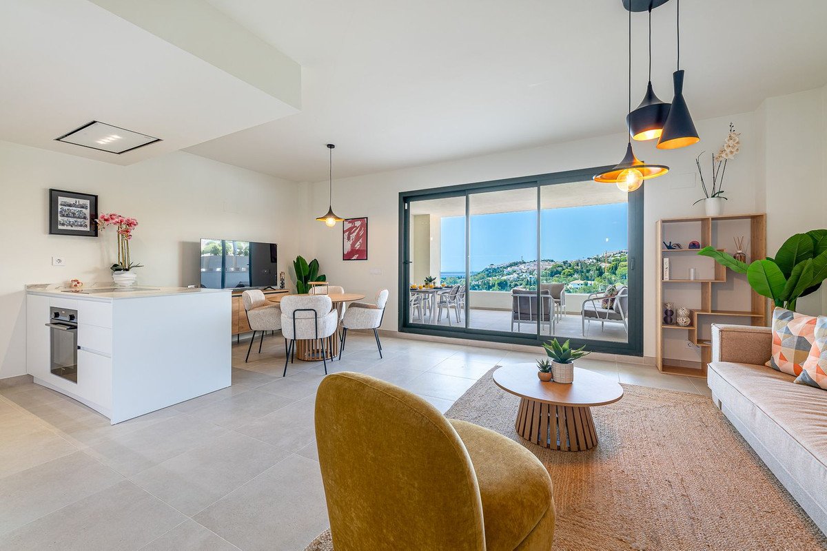 Modern open-plan living, dining, and kitchen area with a balcony offering a sea view and distant hills.