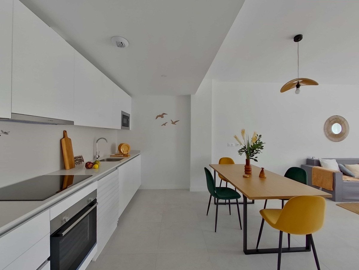 Modern white kitchen with a dining table and chairs, featuring a large window and a sofa in the background.