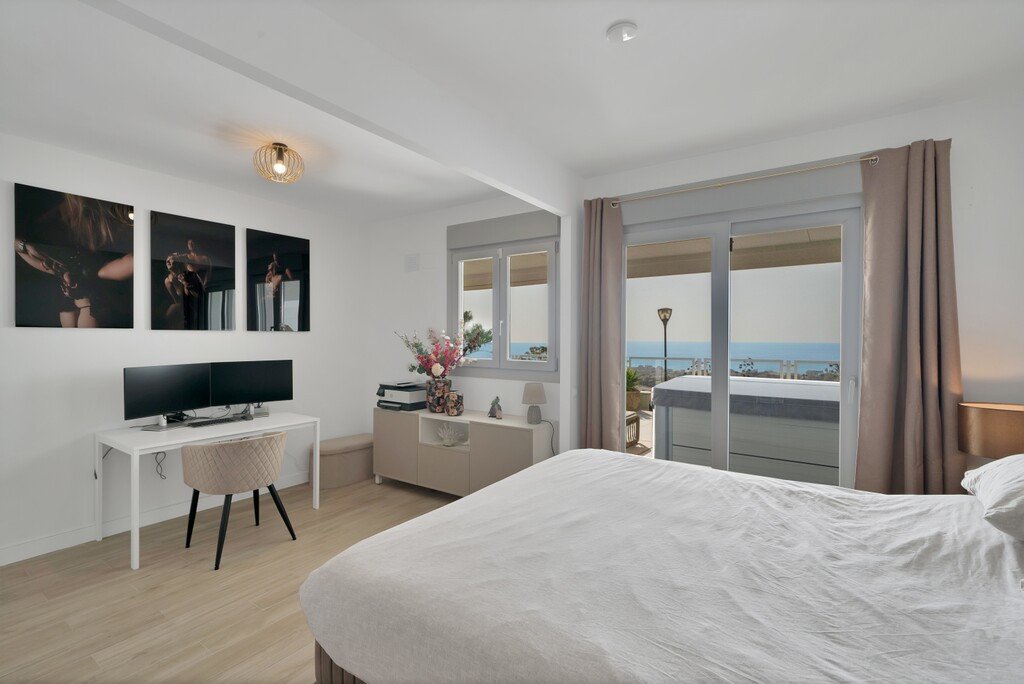 Modern bedroom with dual monitors, artwork, and a balcony overlooking the ocean and hot tub.