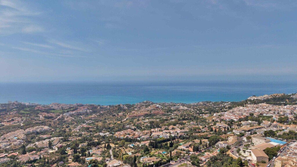 Panoramic aerial view of a coastal town with buildings nestled amongst green hills overlooking the blue ocean under a clear sky.