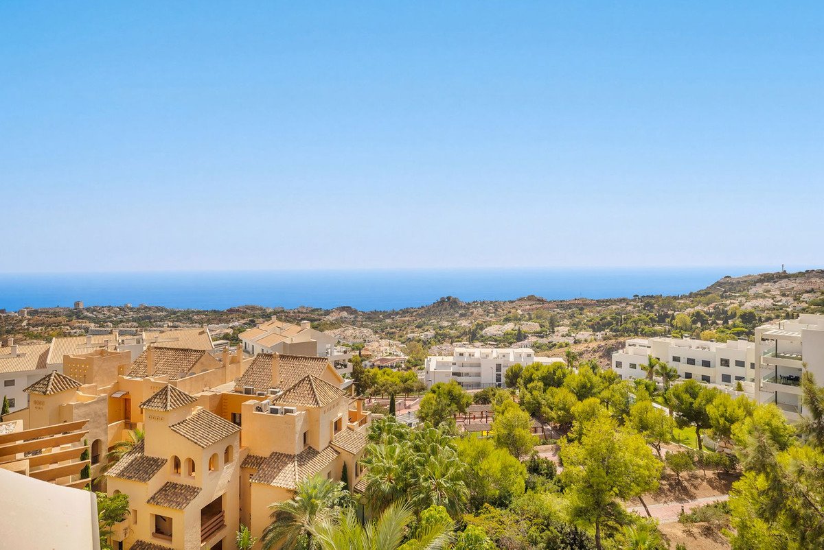 Expansive view from a property showing terracotta-roofed buildings, lush green trees, and the blue sea under a clear sky.